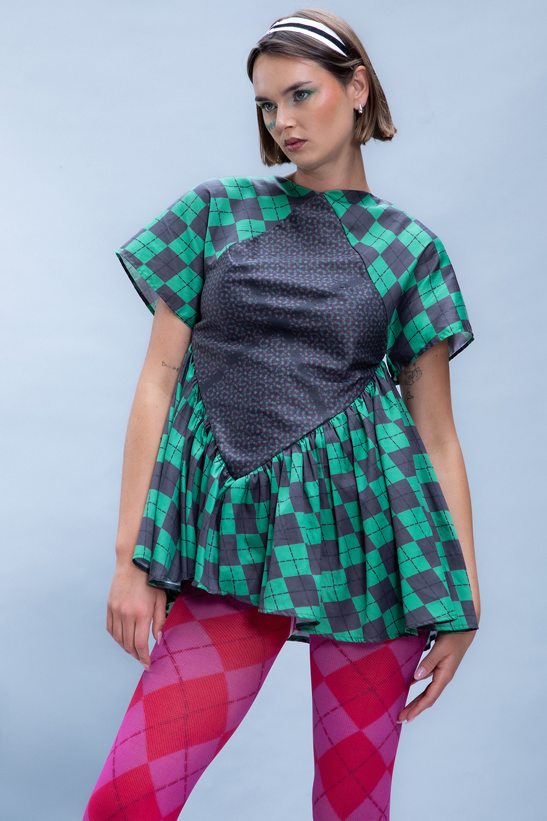 Model wearing a green checkered tunic shot from the knees up posing in contraposto stance against a gray backdrop